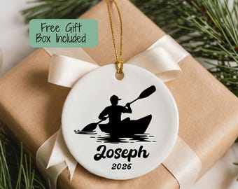 May include: A round white ceramic ornament featuring a black silhouette of a person kayaking. The text Joseph and the year 2026 are printed in black below the graphic. A gold string is attached to the top for hanging purposes.