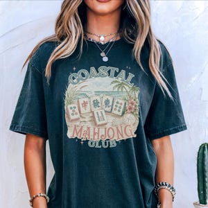 May include: A dark charcoal gray short-sleeve t-shirt featuring a graphic print with the text COASTAL MAHJONG CLUB. The design includes illustrations of mahjong tiles, palm trees, ocean waves, and seashells in muted tones of green, tan, and pink.