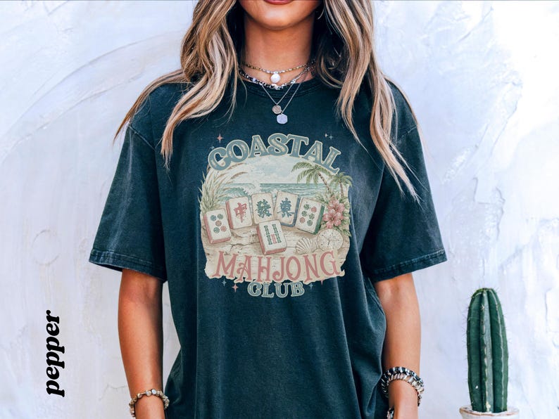 May include: A dark charcoal gray short-sleeve t-shirt featuring a graphic print with the text COASTAL MAHJONG CLUB. The design includes illustrations of mahjong tiles, palm trees, ocean waves, and seashells in muted tones of green, tan, and pink.