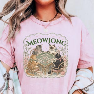 K&ouml;nnte beinhalten: Ein hellrosa T-Shirt mit kurzen &Auml;rmeln und einem Grafikdruck von vier Katzen, die an einem Mahjong-Tisch sitzen. Der Text MEOWJONG ist in Gr&uuml;n &uuml;ber den Katzen gedruckt. Die Illustration enth&auml;lt florale Akzente und Bambusstangen im Rahmen.