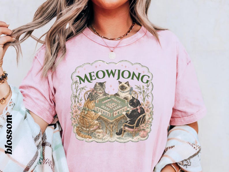 K&ouml;nnte beinhalten: Ein hellrosa T-Shirt mit kurzen &Auml;rmeln und einem Grafikdruck von vier Katzen, die an einem Mahjong-Tisch sitzen. Der Text MEOWJONG ist in Gr&uuml;n &uuml;ber den Katzen gedruckt. Die Illustration enth&auml;lt florale Akzente und Bambusstangen im Rahmen.