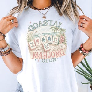 May include: A white t-shirt featuring a graphic print of mahjong tiles on a sandy beach with palm trees and flowers. The text reads Coastal Mahjong Club in a vintage-style font. The design includes small star accents and a compass illustration.