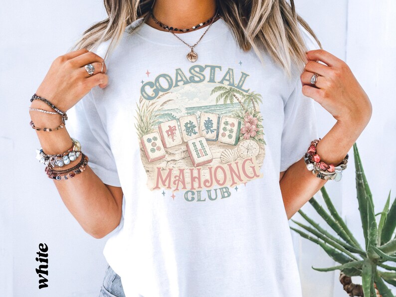 May include: A white t-shirt featuring a graphic print of mahjong tiles on a sandy beach with palm trees and flowers. The text reads Coastal Mahjong Club in a vintage-style font. The design includes small star accents and a compass illustration.