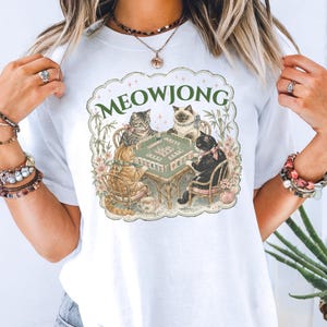 K&ouml;nnte beinhalten: Ein wei&szlig;es T-Shirt mit einem Grafikdruck von vier Katzen, die an einem Tisch sitzen und Mahjong spielen. Der Text MEOWJONG ist in Gr&uuml;n &uuml;ber den Katzen aufgedruckt, umgeben von einem dekorativen, gewellten Rand mit Blumen- und Bambuselementen.