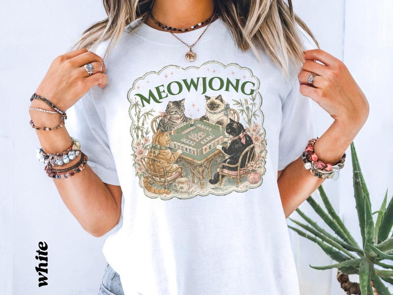 K&ouml;nnte beinhalten: Ein wei&szlig;es T-Shirt mit einem Grafikdruck von vier Katzen, die an einem Tisch sitzen und Mahjong spielen. Der Text MEOWJONG ist in Gr&uuml;n &uuml;ber den Katzen aufgedruckt, umgeben von einem dekorativen, gewellten Rand mit Blumen- und Bambuselementen.