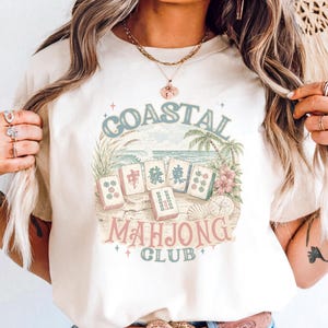 May include: An ivory t-shirt featuring a graphic print of mahjong tiles arranged on a sandy beach with palm trees and hibiscus flowers. The text COASTAL MAHJONG CLUB is printed in a curved layout around the tiles in muted blue and pink tones.