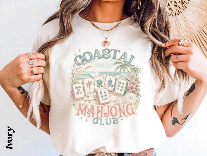 May include: An ivory t-shirt featuring a graphic print of mahjong tiles arranged on a sandy beach with palm trees and hibiscus flowers. The text COASTAL MAHJONG CLUB is printed in a curved layout around the tiles in muted blue and pink tones.