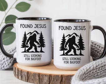 May include: Two white ceramic mugs with black handles and interiors. Each mug features a black graphic of Bigfoot walking between two pine trees with mountains in the background. The text reads FOUND JESUS above and STILL LOOKING FOR BIGFOOT below.