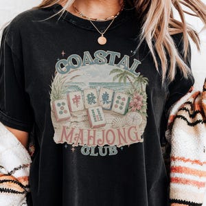 May include: A black short-sleeve t-shirt featuring a graphic print of five mahjong tiles on a sandy beach with palm trees and ocean waves. The text COASTAL MAHJONG CLUB is printed in a vintage-style font around the central illustration.
