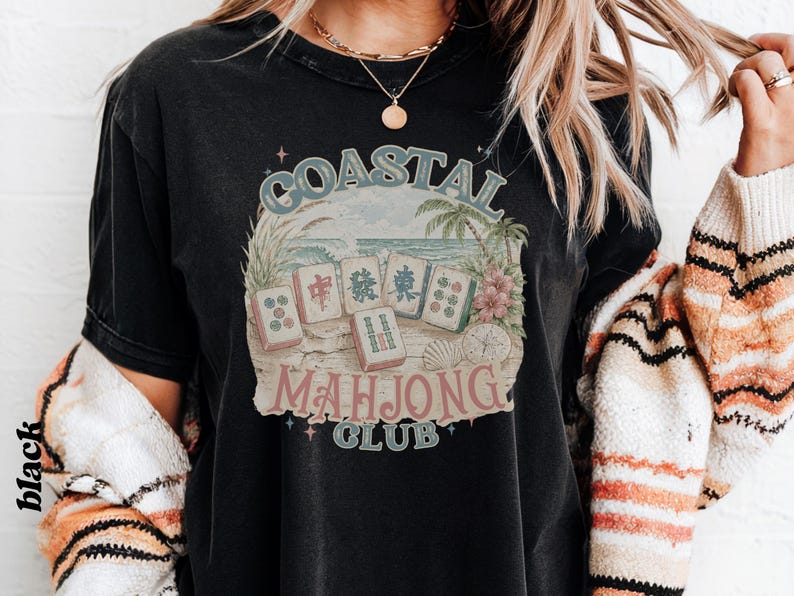 May include: A black short-sleeve t-shirt featuring a graphic print of five mahjong tiles on a sandy beach with palm trees and ocean waves. The text COASTAL MAHJONG CLUB is printed in a vintage-style font around the central illustration.