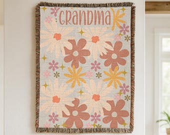 May include: A woven throw blanket with a floral pattern in shades of peach, brown, yellow, and pink. The word "GRANDMA" is displayed in a pink, stylized font. The blanket has a fringed edge and is displayed on a wooden shelf.
