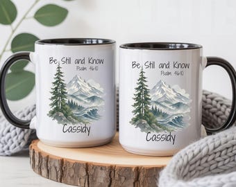 May include: Two white ceramic mugs with black rims and handles, featuring a watercolor mountain and pine tree landscape graphic. The text reads Be Still and Know Psalm 46:10 and the name Cassidy in black script font on the front of each mug.