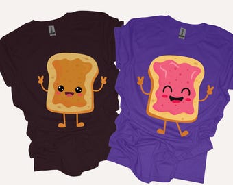 May include: A dark brown t-shirt and a purple t-shirt, each featuring a cartoon slice of toast. The brown shirt shows toast with peanut butter, while the purple shirt shows toast with pink jelly. Both characters have smiling faces and arms making peace signs.
