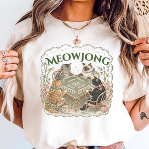 K&ouml;nnte beinhalten: Ein elfenbeinfarbenes T-Shirt mit einem Grafikdruck von vier Katzen, die an einem Mahjong-Tisch sitzen. Der Text MEOWJONG ist in Gr&uuml;n &uuml;ber den Katzen gedruckt. Die Illustration enth&auml;lt Bambusstangen und florale Akzente in einem Rahmen.