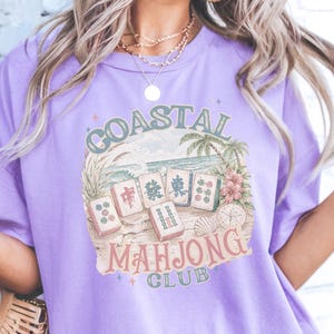 May include: A light purple t-shirt featuring a graphic print with the text COASTAL MAHJONG CLUB. The design includes five mahjong tiles arranged on a sandy beach with palm trees, ocean waves, a seashell, and a sand dollar in muted colors.