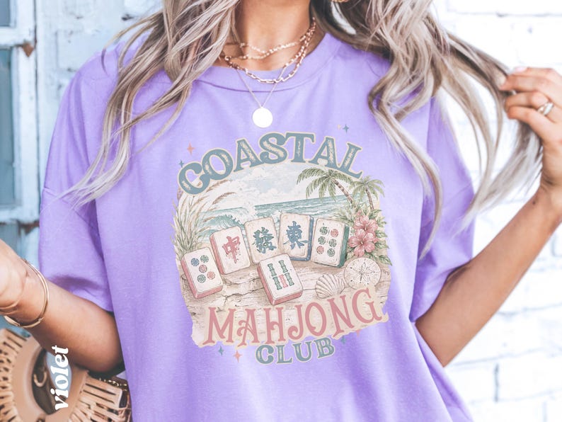 May include: A light purple t-shirt featuring a graphic print with the text COASTAL MAHJONG CLUB. The design includes five mahjong tiles arranged on a sandy beach with palm trees, ocean waves, a seashell, and a sand dollar in muted colors.