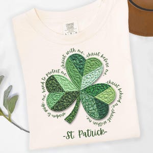 May include: Ivory-colored t-shirt featuring a detailed green shamrock design. The shamrock is surrounded by text and the words "St. Patrick" are below. The shirt is made of a soft material.