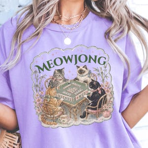 K&ouml;nnte beinhalten: Ein hellviolettes T-Shirt mit einem Grafikdruck von vier Katzen, die an einem Tisch sitzen und Mahjong spielen. Der Text MEOWJONG ist in Gr&uuml;n &uuml;ber den Katzen aufgedruckt, umgeben von floralen Akzenten und Bambusstangen in einem Rahmen.