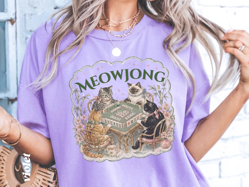 K&ouml;nnte beinhalten: Ein hellviolettes T-Shirt mit einem Grafikdruck von vier Katzen, die an einem Tisch sitzen und Mahjong spielen. Der Text MEOWJONG ist in Gr&uuml;n &uuml;ber den Katzen aufgedruckt, umgeben von floralen Akzenten und Bambusstangen in einem Rahmen.