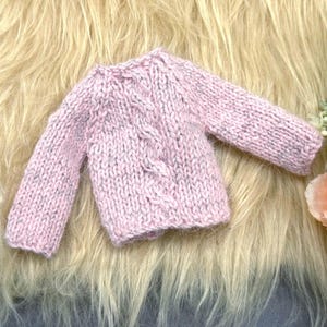Può includere: Un maglione a maniche lunghe rosa chiaro con un motivo a trecce verticali al centro. Il capo è realizzato in filato di lana grossa con sottili macchie di colore più scuro, scollo rotondo e polsini a coste sulle maniche.