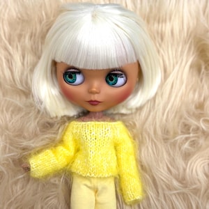 Puede incluir: Una muñeca Blythe con un corte de pelo bob blanco y grandes ojos turquesa. La muñeca lleva un suéter amarillo con hombros descubiertos y pantalones a juego. La piel de la muñeca es de color canela claro, y el fondo es un material esponjoso color crema.