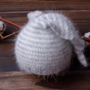 May include: A soft, gray knitted hat with horizontal stripes and a long, pointed tip. The hat is fuzzy and rests on a wooden surface, with cotton branches in the background. The hat is likely for a baby.
