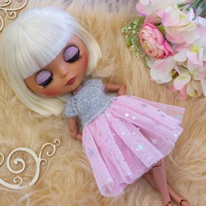 May include: A doll with blonde hair and closed eyes, wearing a gray top and a pink tulle skirt with silver star accents. The doll is lying on a fluffy, light brown surface, with flowers and wooden decorative elements nearby.