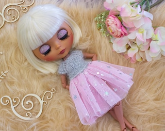 May include: A doll with blonde hair and closed eyes, wearing a gray top and a pink tulle skirt with silver star accents. The doll is lying on a fluffy, light brown surface, with flowers and wooden decorative elements nearby.