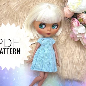 May include: A doll with a light blue knitted dress. The doll has white hair and large, expressive eyes. The image includes the text "PDF PATTERN" in a white starburst shape. The background features a fluffy, light-colored surface and pink and white flowers.