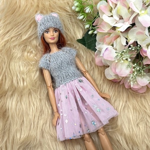 May include: A doll wearing a gray knit hat with mouse ears, a gray knit top, and a pink tulle skirt with silver star-shaped sequins. The doll is lying on a fluffy, light brown surface, with pink and white flowers in the background.