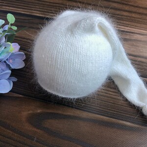 May include: A soft, white knitted baby hat with a long, pointed tail. The hat is made of a fuzzy, textured yarn and is designed to fit a newborn. The hat is sitting on a dark brown wooden surface.