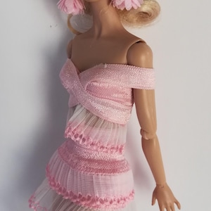 May include: A fashion doll with blonde hair styled in an updo, wearing pink tassel earrings and a light pink off-the-shoulder dress. The dress features a crisscross bodice, layered pleated fabric, and decorative scalloped trim along the hem.