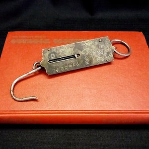 May include: An antique metal hanging scale with a hook and ring, marked "NEW YORK" and "20 LBS", rests on a red book. The scale has a sliding indicator and a measurement scale from 0 to 20.