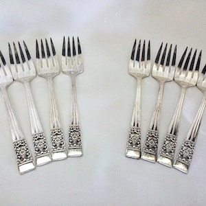 May include: A collection of silver forks with ornate floral designs on the handles. The forks have four tines and are arranged on a white surface. The silverware is likely for formal dining.