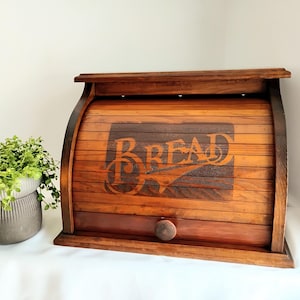 May include: A dark brown wooden bread box with a roll-top door featuring the word BREAD in a decorative font. The box has a small round knob on the front panel and a flat top surface. A small green plant in a gray pot sits to the left.