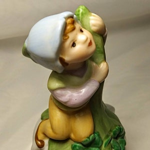 May include: A porcelain figurine of a boy wearing a blue hat and green shirt, climbing a green vine. The boy has brown hair and is holding onto the vine with both hands. The figurine is sitting on a green base with shamrock leaves.