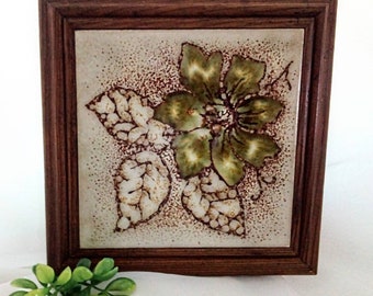 May include: A framed ceramic tile with a green and white floral design. The tile has a brown speckled background and is framed in a dark wood frame.