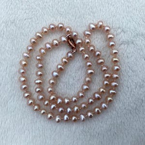 Puede incluir: Un collar compuesto por numerosas perlas de agua dulce pequeñas, de color rosa claro y forma irregular, ensartadas en un hilo continuo. Las perlas presentan un brillo suave y lustroso y están aseguradas con un pequeño cierre metálico arriba.