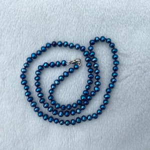 Könnte beinhalten: Eine Halskette bestehend aus zahlreichen kleinen, runden, dunkelblauen Perlen mit metallischem Glanz. Die Perlen sind auf einem durchgehenden Strang aufgereiht und werden durch einen silberfarbenen Karabinerverschluss aus Metall zusammengehalten.
