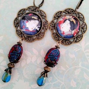 Op de afbeelding: Een paar oorhangers met een geisha-afbeelding in een bronzen filigraan bloem. De oorbellen hebben rood-blauwe kralen met patronen en druppelvormige blauwe glazen druppels. De oorbellen zijn ontworpen met een oorhaaksluiting.