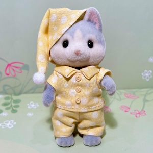 May include: A plush toy cat wearing yellow pajamas with white polka dots and a matching nightcap. The cat has gray fur and black eyes. The pajamas include a button-down shirt and pants. The nightcap has a white pom-pom.