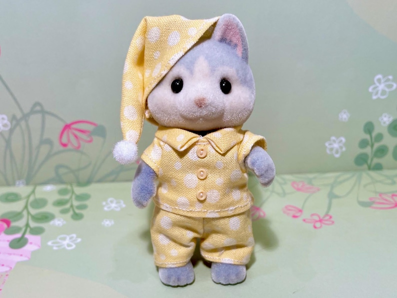 May include: A plush toy cat wearing yellow pajamas with white polka dots and a matching nightcap. The cat has gray fur and black eyes. The pajamas include a button-down shirt and pants. The nightcap has a white pom-pom.