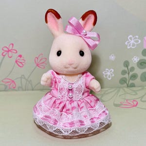 Può includere: Una statuetta di coniglio floccato con pelliccia color crema e punte delle orecchie marroni. Il coniglio indossa un vestito rosa a motivi con bordo in pizzo bianco, due piccoli bottoni perlati sul corpetto e un fiocco rosa con perla.