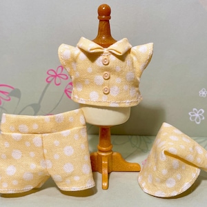May include: A miniature yellow and white polka dot pajama set, including a collared shirt, shorts, and a nightcap with a white pom-pom. The set is displayed on a small mannequin, showcasing the details of the sleepwear.