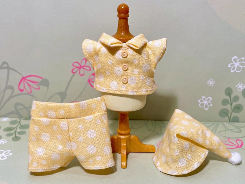 May include: A miniature yellow and white polka dot pajama set, including a collared shirt, shorts, and a nightcap with a white pom-pom. The set is displayed on a small mannequin, showcasing the details of the sleepwear.