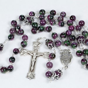 May include: A rosary with round beads in shades of purple and green, strung on a silver chain. The rosary features a silver crucifix and a silver pendant with a religious figure.