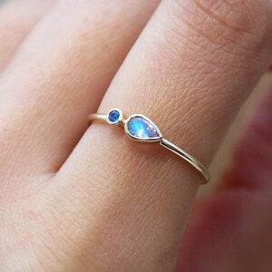 Pode incluir: Um delicado anel de ouro com duas pedras preciosas. Uma &eacute; uma pequena pedra azul redonda, e a outra &eacute; uma pedra iridescente em forma de p&ecirc;ra. O anel &eacute; mostrado em um dedo, destacando seu design minimalista e seu apelo elegante.