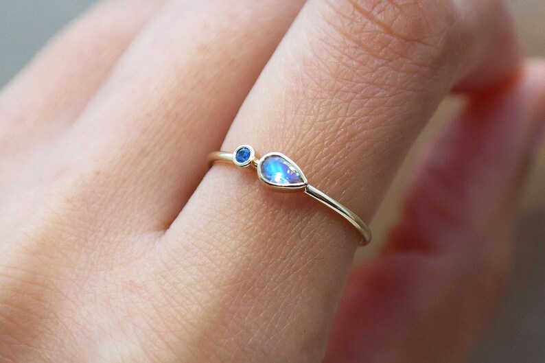 Pode incluir: Um delicado anel de ouro com duas pedras preciosas. Uma &eacute; uma pequena pedra azul redonda, e a outra &eacute; uma pedra iridescente em forma de p&ecirc;ra. O anel &eacute; mostrado em um dedo, destacando seu design minimalista e seu apelo elegante.