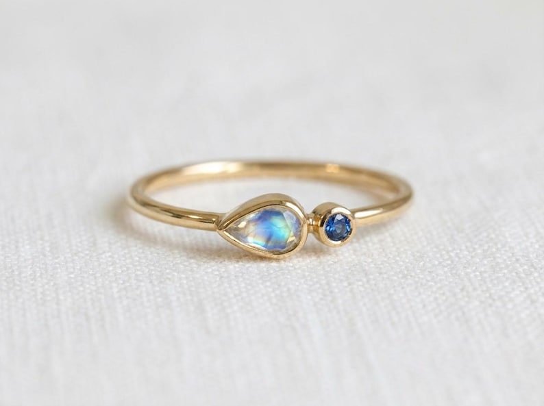 Pode incluir: Um delicado anel de ouro com uma pedra iridescente em forma de p&ecirc;ra e uma pequena pedra azul redonda. O anel tem uma banda fina e polida. As pedras est&atilde;o pr&oacute;ximas, criando um design minimalista e elegante. Uma joia.