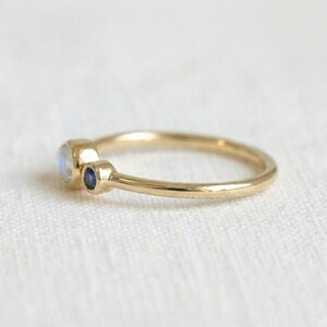 Pode incluir: Um delicado anel de ouro com uma pedra da lua redonda e iridescente e uma pequena safira azul escura. O anel tem uma banda fina e um design minimalista. As pedras preciosas s&atilde;o definidas em uma configura&ccedil;&atilde;o de moldura.